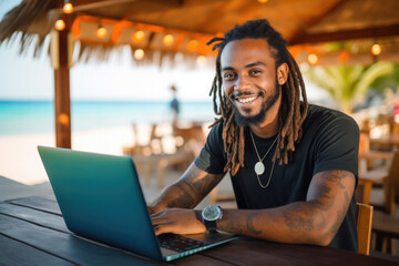 Smiling handsome hipster black man with dreadlocks  sitting and working on his laptop in the beach bar