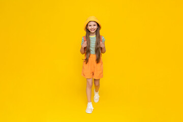 A beautiful little girl in full-length shorts and a panama hat is smiling happily. A child with a backpack is enjoying the summer. Yellow isolated background.
