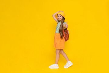 A beautiful little girl in full - length shorts and a panama hat smiles happily and gives a thumbs up . A child with a backpack is enjoying the summer. Yellow isolated background.