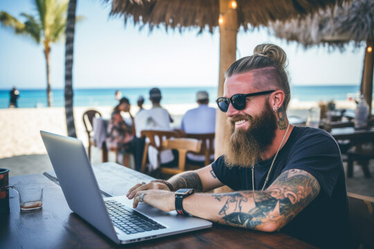 Smiling handsome hipster man with tattoos sitting and working on his laptop in the beach bar