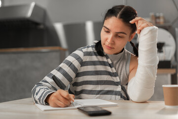 Beautiful girl with broken arm is making notes in cafeteria. Freelance and remote work. Modern...
