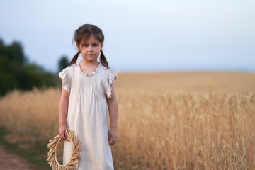 A little girl in a linen dress with a wreath of ears in her hand on the field road of a wheat field.