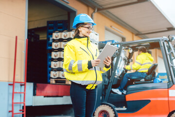 woman in industrial gear standing in front of a forklift transporting goods from a warehouse