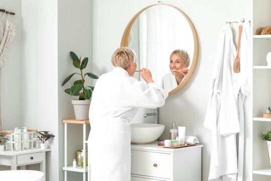 Mature woman brushing teeth near mirror in bathroom