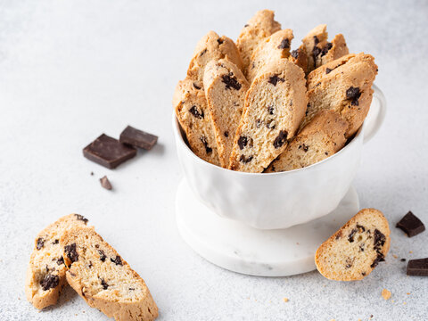 Cantucci with chocolate pieces in white cup. Italian classic biscotti with dark chocolate chips and orange zest. Close up view. Homemade crunchy biscuits. Copy space. White background.