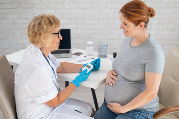 An elderly female doctor checks the blood sugar level of a pregnant red-haired woman. Gestational...