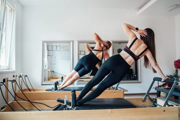 Foto op Canvas Two smiling fit-shaped females doing side plank with raised arm static core muscles exercise using pilates reformer machine in the sport athletic gym with a mirror wall. Active people training concept © Soloviova Liudmyla