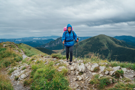 Young smiling hiker woman dressed rain suit walking by rocky Slovakian Mala Fatra mountain range using tracking poles with backpack. Active lifestyle concept image