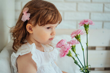child girl smells a flower near the window. a girl in a white T-shirt, with a pink hairpin and brown hair. A child looks out the window waiting for spring