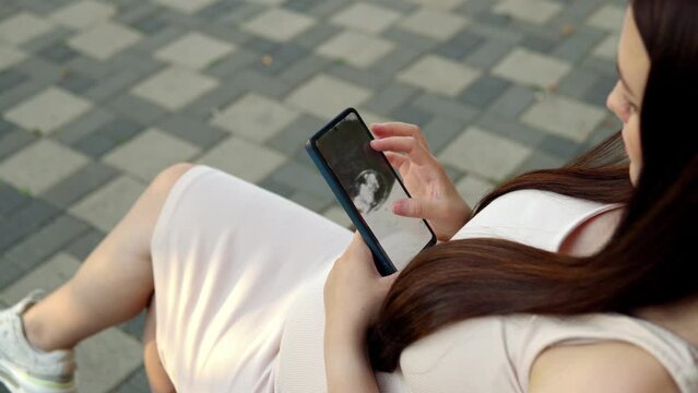 In a bustling public place, a pregnant young woman sits on a bench, deeply engrossed in examining an ultrasound photo of her baby on her smartphone.