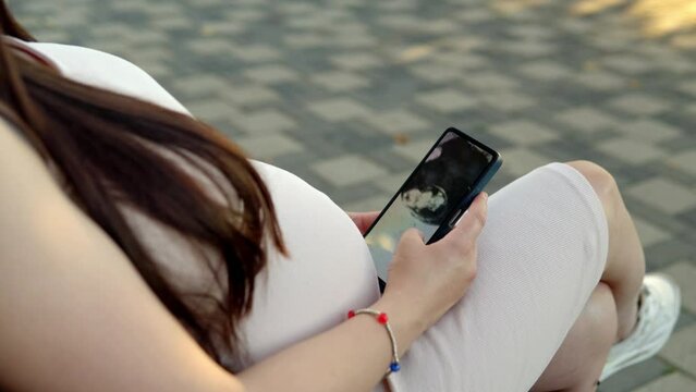 In a bustling public place, a pregnant young woman sits on a bench, deeply engrossed in examining an ultrasound photo of her baby on her smartphone.