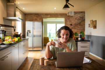 Middle aged mixed ethnicity woman drinking tea while working and using her laptop in the kitchen at home