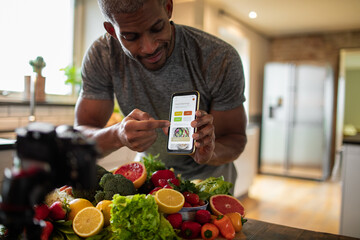 Middle aged african american male food nutritionist and blogger promoting and showing his dieting app to his customers and clients while recording in the kitchen with organic fruits and vegetables