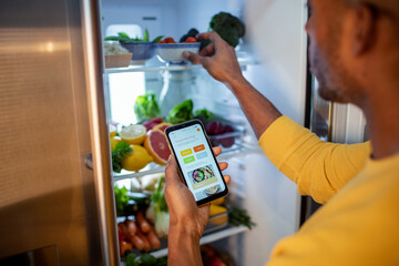 Middle aged african american man using a dieting app while looking through his fridge in the kitchen at home