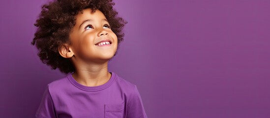 Adorable curly haired boy holds hands looks up dreamily and smiles in a casual t shirt on purple background