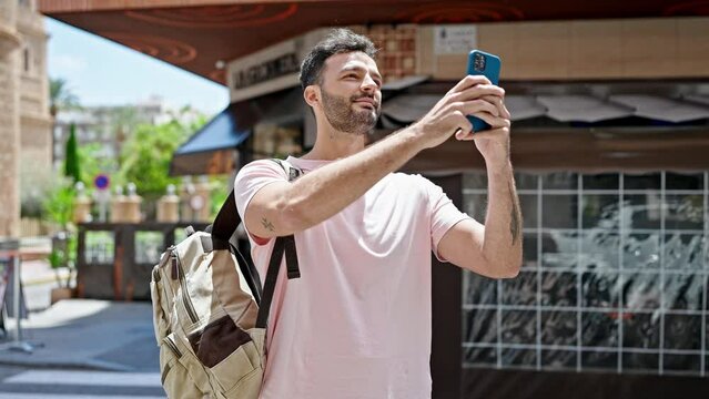 Young hispanic man recording video by smartphone wearing backpack at coffee shop terrace