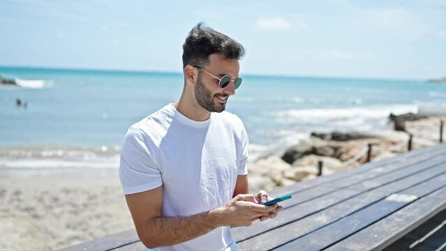 Young hispanic man using smartphone smiling at seaside