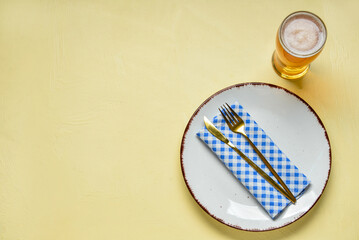 Empty plate with cutlery, blue checkered napkin and mug of beer on yellow background