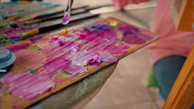 A close-up of a girl artist sitting near an easel and taking paint from a palette with a brush the concept of love of fine art 