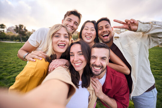 Group of young cheerful multi-ethnic friends taking selfie with mobile phone outdoor. Happy people together smiling and enjoying free time in park. Positive and fun youth relationships in community.