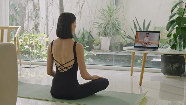 Woman sitting in butterfly pose on exercise mat at home, watching video workout on laptop and listening to fitness coach before stretching