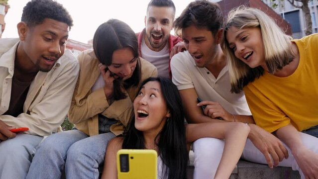 Group young people surprised watching something on phone. Multiracial friends sitting outdoor using mobile together. Concept of friendship social networks and technology addiction in new generations.