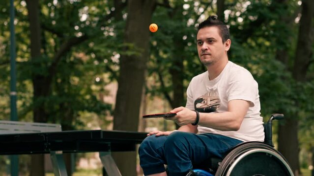A man in a wheelchair concentrates on hitting an orange ball with a tennis racket after playing ping pong in a city park