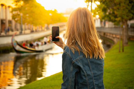 40 years old blonde tourist woman with mobile phone taking a photo front sea canal with Moliceiro boat at golden sunset Aveiro, Portugal 