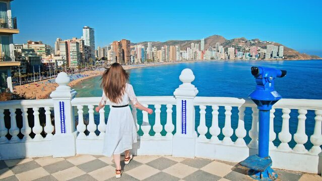 A beautiful girl in a dress looking at a scenic view of Spanish Beach and the Sea. A woman enjoying panoramic views of Platja de Llevant and Cala del Mal Pas Beach from above in Benidorm, Alicante
