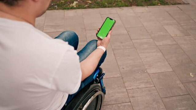 A man in a wheelchair holds a phone with an open green screen in a city park view from behind