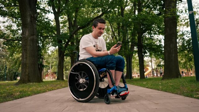 young man in a wheelchair looks intently at the phone emotionally reads the news or communicates in the messenger in the city park technology mobility