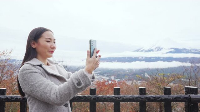 Asian woman using mobile phone taking selfie or vlogging with mt Fuji covered in snow with beautiful red maple tree leaves falling during travel nature lake Kawaguchi, Japan in autumn holiday vacation