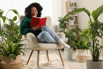 Relaxing atmosphere. Happy woman reading book on armchair surrounded by beautiful houseplants in...