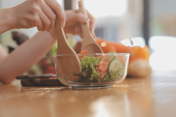 Delicious fruit and vegetables on a table and woman cooking. Housewife is cutting green cucumbers on a wooden board for making fresh salad in the kitchen.