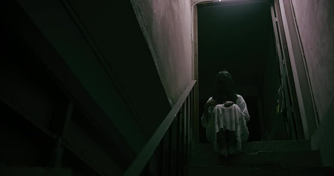 Horror scene of a mysterious Scary Asian ghost woman creepy have hair covering the face sitting on staircase at abandoned house with background dark scene movie at night, festival Halloween concept