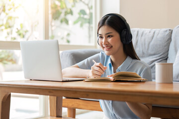 young woman working on laptop and taking notes while listening to lecture at home