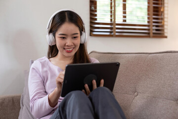 Happy young asian girl in headphones is using a digital tablet and smiling while living at home.