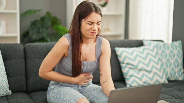 Young beautiful hispanic woman suffering for stomach ache using laptop at home