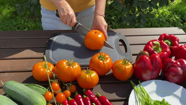 woman slicing yellow tomato with knife on wooden table in summer garden, garden-grown vegetables red tomato, yellow tomato, red bell pepper, cucumber, red hot pepper, parsley