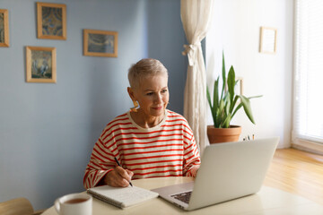 Stylish senior woman in striped shirt studying finances and investments online, using laptop, watching tutorial or podcast and making notes. Elderly people, technology and distant learning