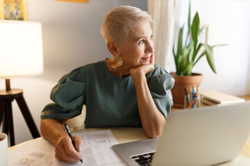 Happy thoughtful senior businesswoman in elegant green dress sitting at table in front of laptop looking through window with smile, putting hand under chin, signing contract, working with documents - obrazy, fototapety, plakaty
