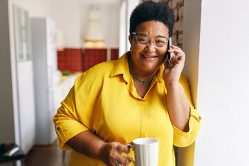 appy cheerful black senior plus size female with short haircut and eyeglasses having coffee break holding white cup while talking on phone with her friend, joking discussing funny situation - 642833916