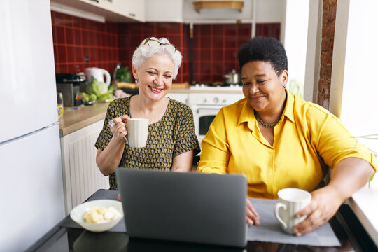 Indoor image of caucasian senior gray-haired woman sitting next to her female best friend of african ethnicity at kitchen table drinking tea and watching comic show on laptop, having fun and rest