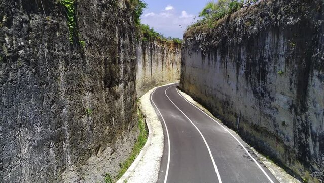 A drone slowly flew through a narrow gap between two cliffs in the Pandawa Beach area, with a winding asphalt road beneath it leading to the Indian Ocean. Aerial cinematic footage.