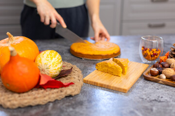 Obraz na płótnie Canvas A woman cuts a cake on the kitchen table and puts it on a wooden board. Delicious baked dessert made from flour and pumpkin. Orange pie. Pie, pumpkins and nuts. Autumn dessert