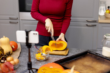 The girl cuts an orange pumpkin and shoots the process on the phone. The blogger broadcasts live or records the lesson on the phone. Cooking in the kitchen.