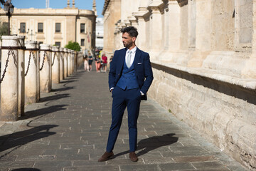 Handsome young man with beard and dressed in blue suit and tie, walks by the cathedral of seville in spain. The man is an executive who is in the city on a business trip.