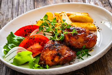 Roasted chicken thighs with French fries and fresh vegetables on wooden table
