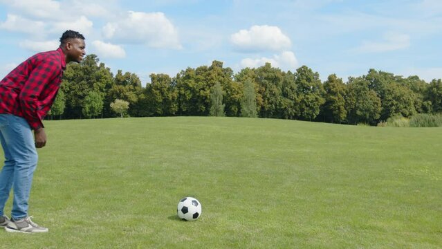 Carefree attractive African man in casual clothes performing penalty kick, kicking soccer ball and celebrating scoring goal with winning gesture while playing football game on field.