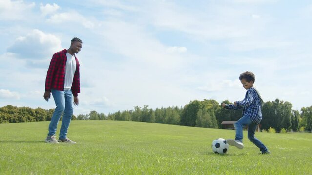 Side view of cheerful united African American family enjoying playing football game on green field, passing soccer ball , training and practicing football skills while spending leisure in nature.